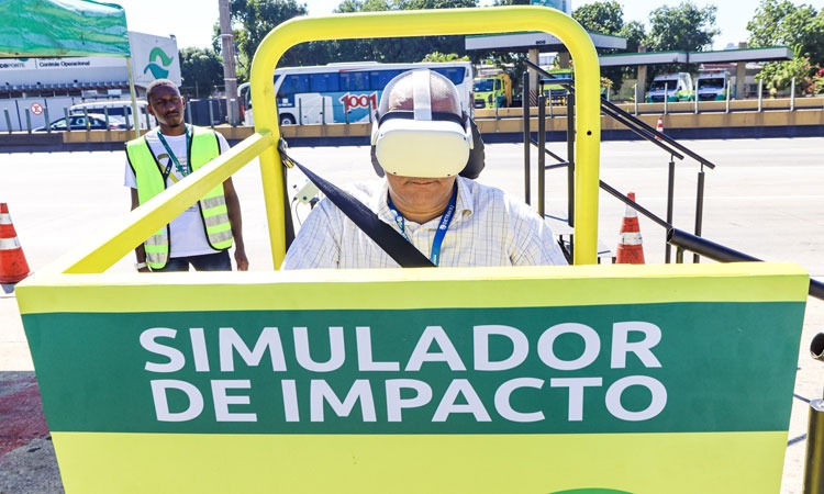 Homem, observado por agente do Detran, participa de atividade com óculos do Detran que simulam reações ao dirigir sob o efeito de com álcool, drogas e sono, dentro de estrutura com mensagem "simulador de impacto".