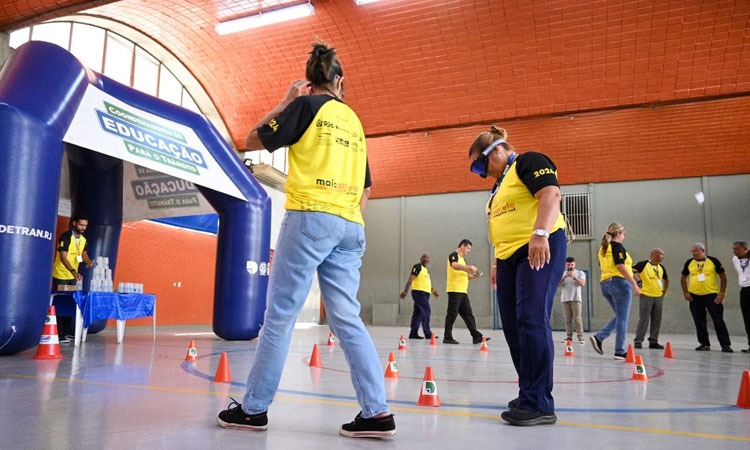 Mulher participa de atividade com óculos do Detran que simulam reações ao dirigir sob o efeito de com álcool, drogas e sono. Ao fundo outras pessoas participam de outra atividade
