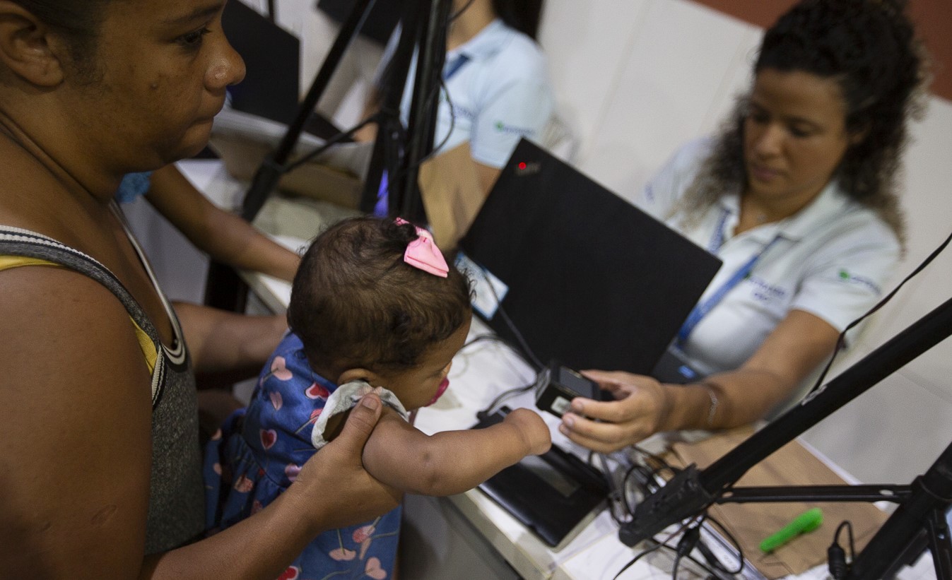 Funcionária do Detran atende uma mulher com seu bebê no colo para tirar a primeira identidade