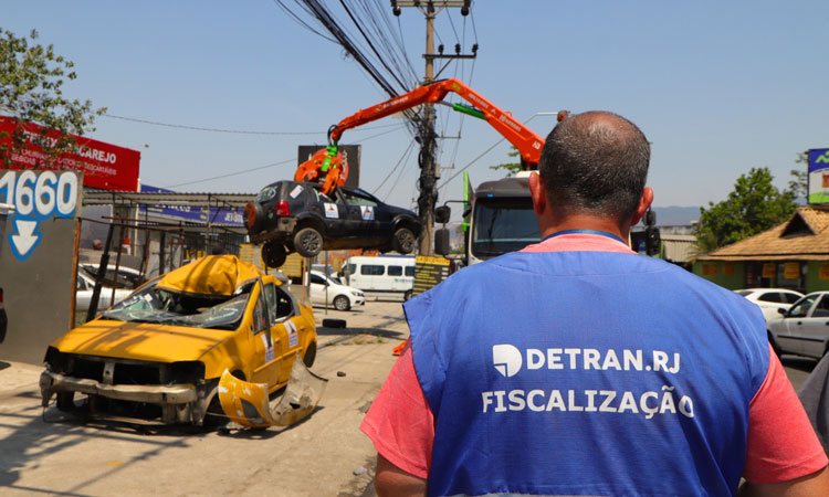 agente de fiscalização do Detran observa carro suspenso por guindaste em ferro-velho