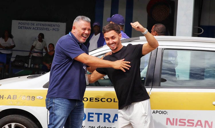 Homem com deficiência comemora aprovação de prova prática de direção junto ao instrutor e em frente a carro adaptado.